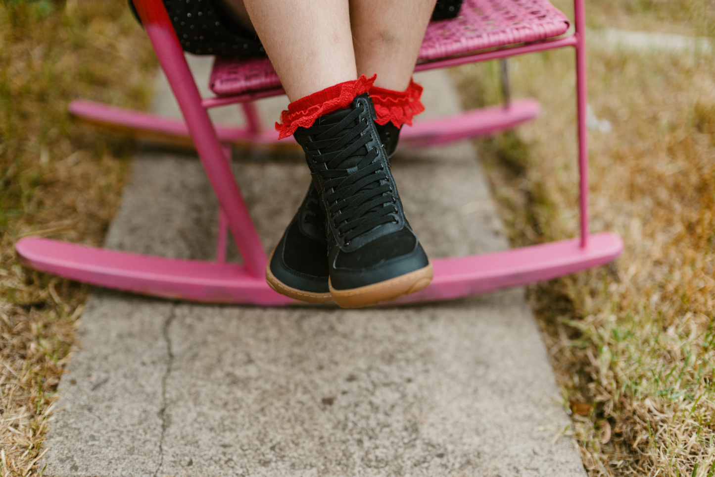 Front view of a child in red frilly socks, sitting on a pink chair, wearing black foot-shaped vegan SNUGG boots by PaperKrane. 