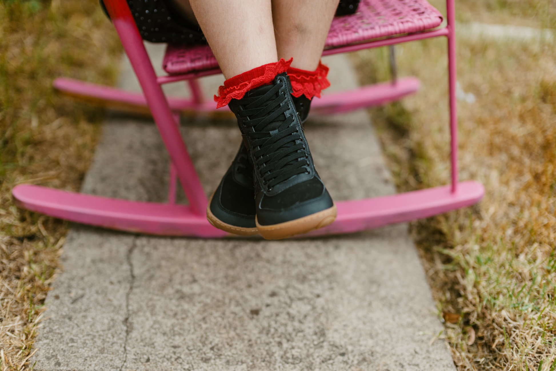 Front view of a child in red frilly socks, sitting on a pink chair, wearing black foot-shaped vegan SNUGG boots by PaperKrane. 