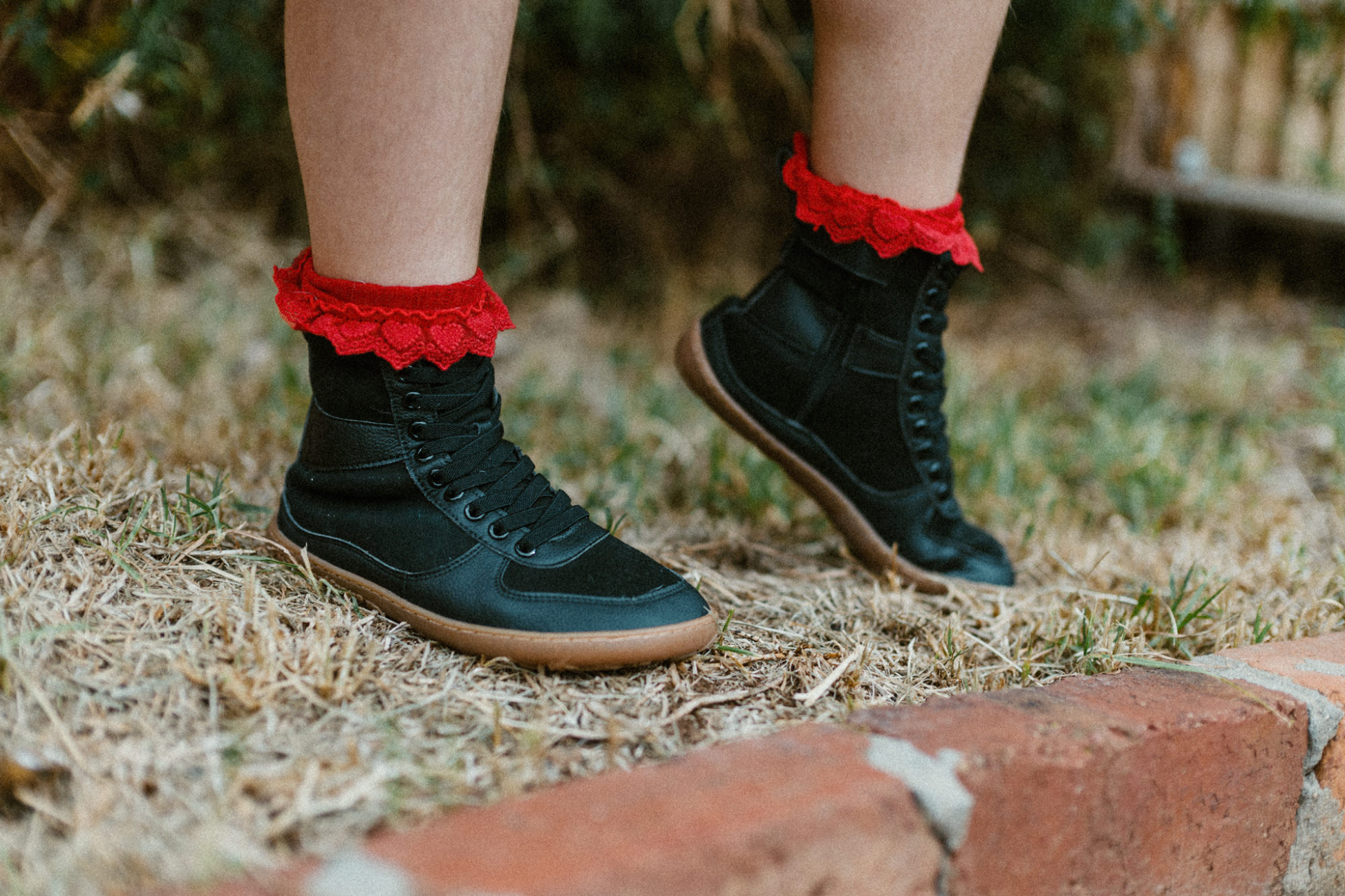 Child standing on grass is wearing foot-shaped SNUGG boots by PaperKrane. The boots are barefoot, and vegan friendly. They're very lightweight and the child is wearing red frilly socks that stick above the top of the tall boots. The laces are elastic. 
