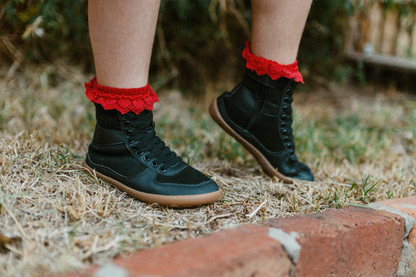Child standing on grass is wearing foot-shaped SNUGG boots by PaperKrane. The boots are barefoot, and vegan friendly. They're very lightweight and the child is wearing red frilly socks that stick above the top of the tall boots. The laces are elastic. 