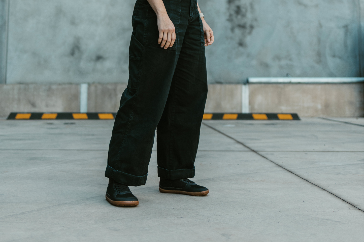 An adult wears PaperKrane vegan SNUGG boots on a concrete background. They're wearing black pants with the black boots. 