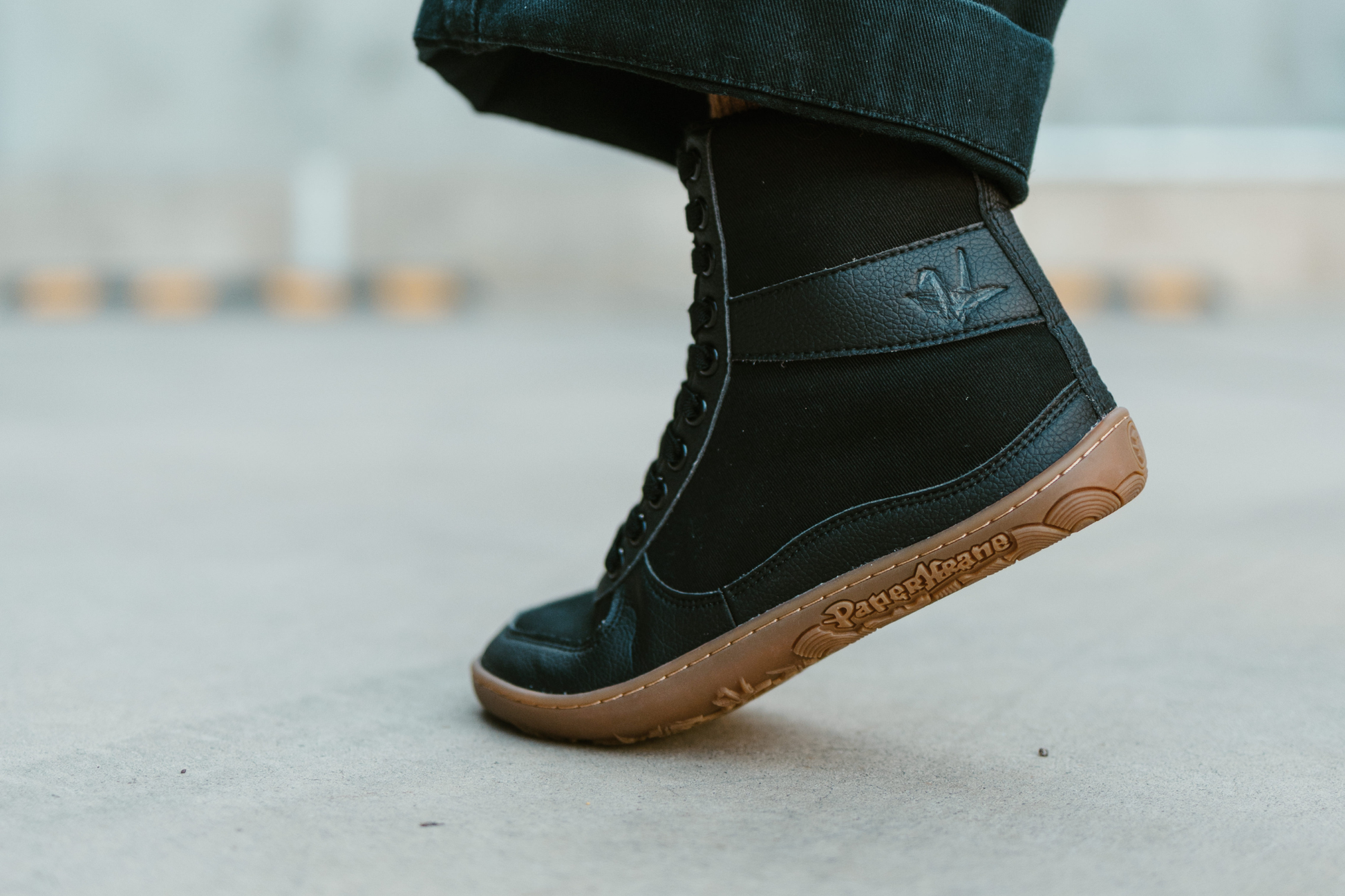 Tip toes! A PaperKrane barefoot SNUGG boot worn by an adult on concrete is raised up on their toe. The boots are black with gum sole and elastic laces. 