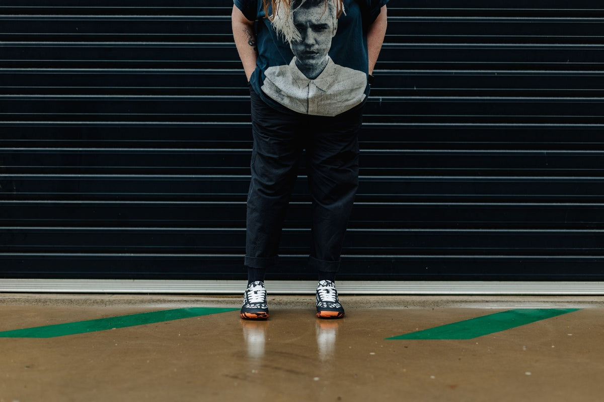 A person is standing on wet concrete in a pair of PaperKrane barefoot sneakers. These are available in JNR and ADULT sizes. The soles are orange and black stripe with white ghosts printed on a black upper. The adult sizes have flat white cotton laces, and the kids pairs up to EU 36 have elastic laces. 