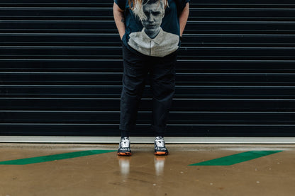 A person is standing on wet concrete in a pair of PaperKrane barefoot sneakers. These are available in JNR and ADULT sizes. The soles are orange and black stripe with white ghosts printed on a black upper. The adult sizes have flat white cotton laces, and the kids pairs up to EU 36 have elastic laces. 