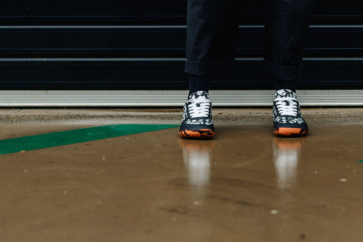 A person wearing black pants and socks is standing on wet concrete in a pair of PaperKrane foot-shaped sneakers with orange and black tiger striped soles, and white ghosts printed on the uppers. They have wide white cotton laces in them. 