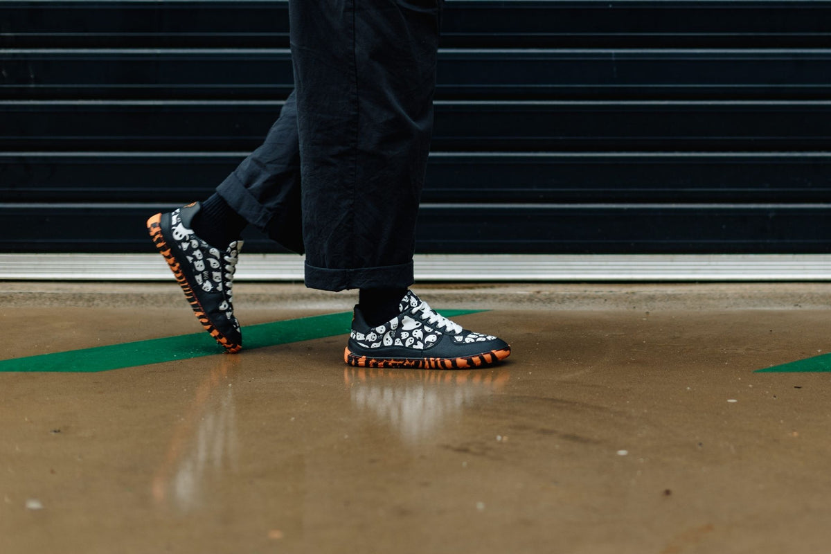 A person wearing black pants and socks is walking on wet concrete in a pair of PaperKrane barefoot sneakers with orange and black tiger striped soles, and white ghosts printed on the uppers. 