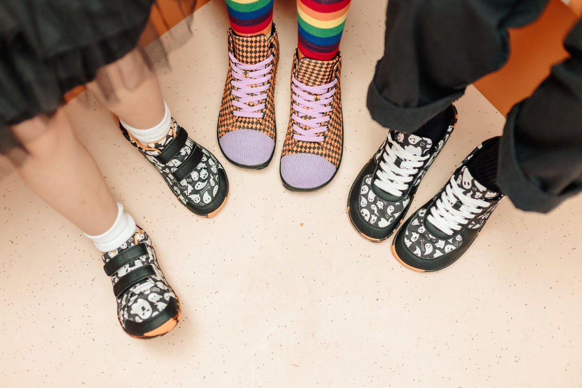 Three pairs of PaperKrane foot-shaped shoes can be seen from above. All have some orange features, the kids ghost print velcros are on the left. A pair of Tall Boots with orange argyle print, purple corduroy on the toes and purple elastic laces are in the middle, worn by someone in rainbow socks, and on the right are the adult ghost print sneakers on a grown up. 