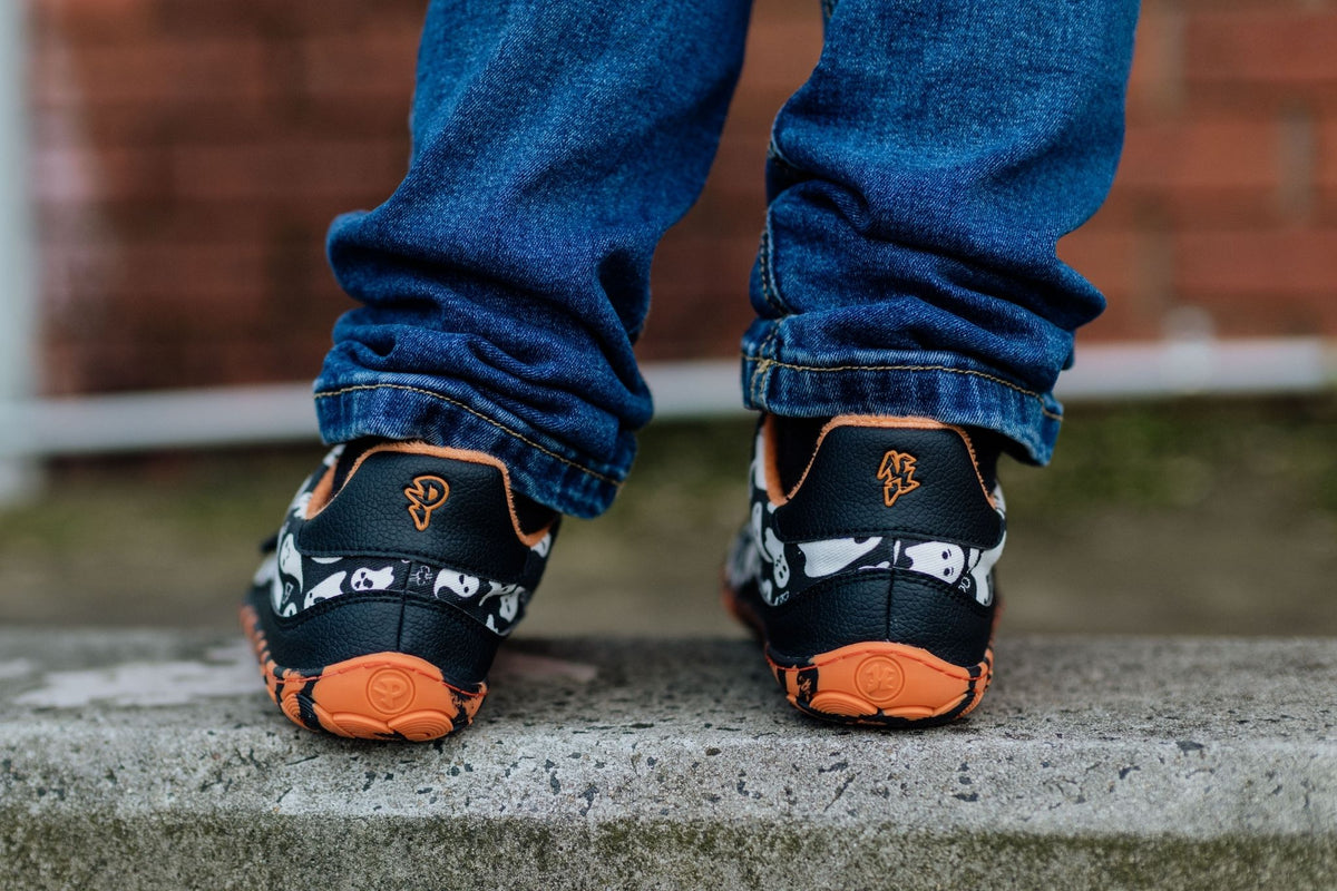 A close-up of a person's feet wearing black and orange ghost print sneakers from PaperKrane, paired with blue jeans, standing on a concrete surface with a blurred brick wall in the background.