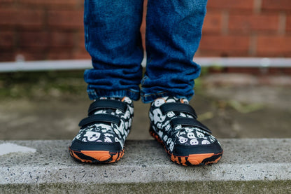 A close-up of a child's feet wearing black and white ghost patterned barefoot sneakers with orange and black striped soles, paired with blue jeans, standing on a concrete ledge. The velcro makes this style super easy for kids to get on. 
