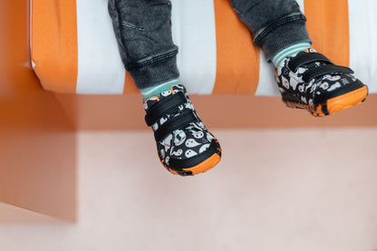A close-up of a child's feet wearing black and white ghost-patterned toddler velcro shoes with orange tiger soles, sitting on an orange and white striped bench.
