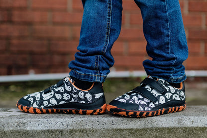 A person wearing black and white ghost-patterned velcro sneakers from PaperKrane with bright orange and black striped soles, paired with blue jeans, standing on a concrete ledge with a brick wall in the background.