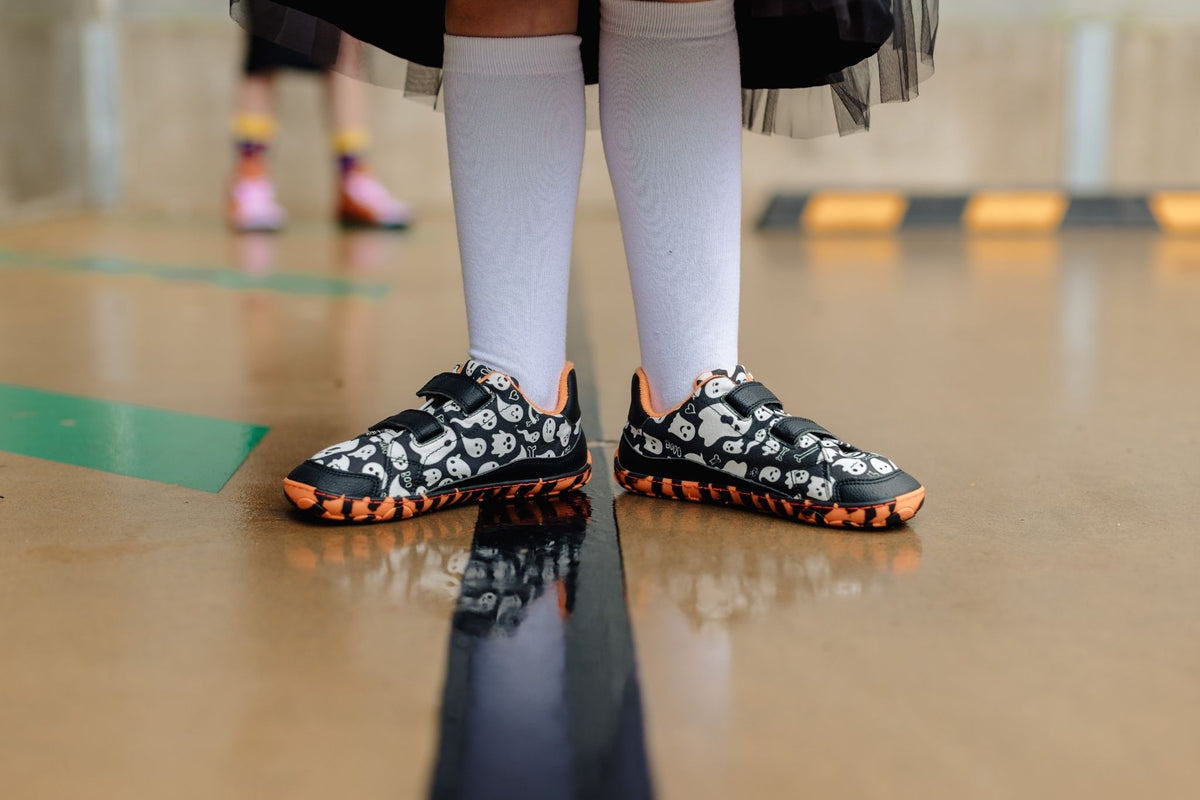 A close-up of a person's legs wearing black and white ghost patterned flat barefoot shoes with bright orange and black striped soles, paired with white knee-high socks, standing on a wet concrete floor.