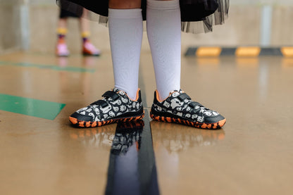 A close-up of a person's legs wearing black and white ghost patterned flat barefoot shoes with bright orange and black striped soles, paired with white knee-high socks, standing on a wet concrete floor.