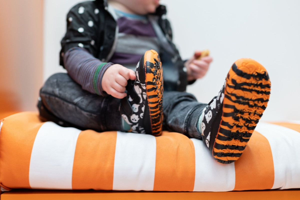 A child sitting on an orange and white striped cushion, wearing black and orange tiger-print slip-on velcro shoes with a ghost pattern on the uppers.