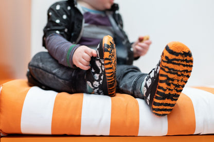 A child sitting on an orange and white striped cushion, wearing black and orange tiger-print slip-on velcro shoes with a ghost pattern on the uppers.