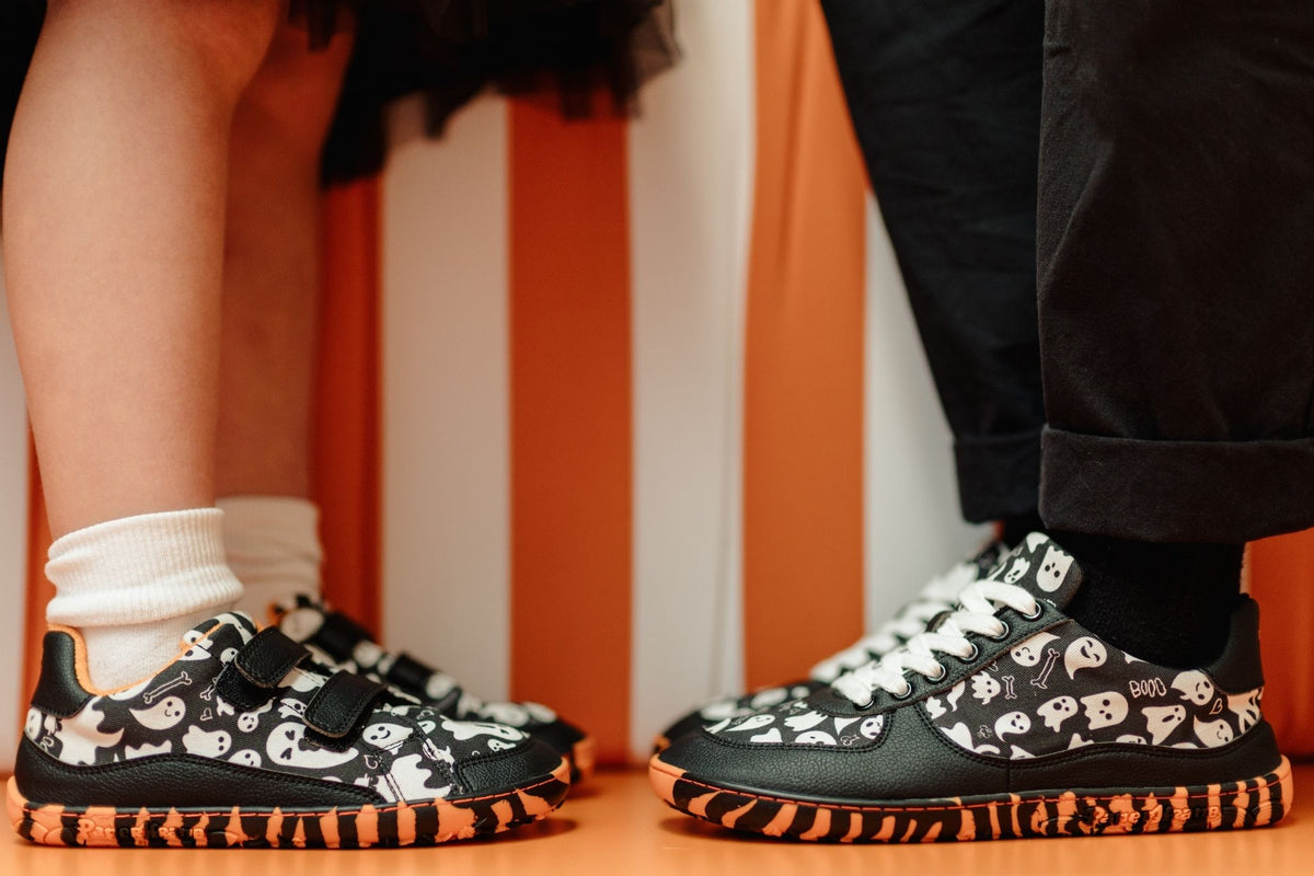 A child and adult are standing toe-to-toe in matching pairs of vegan PaperKrane sneakers. The child has on a pair of the ghost prints shoes with orange and black striped soles, with velcro straps, and the adult is wearing the matching ghost print sneaker which has white cotton laces. 