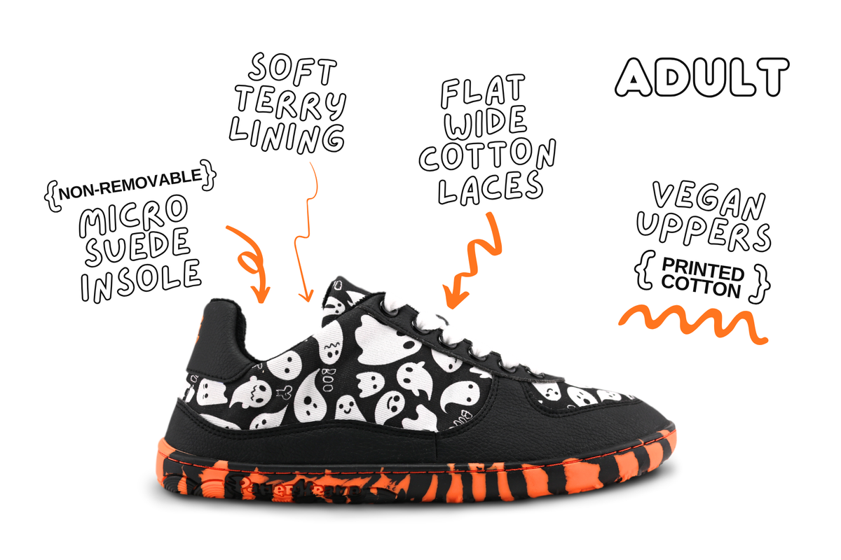 A pair of PaperKrane barefoot sneakers with orange and black striped rubber soles, black vegan leather, and white ghosts printed on cotton. They're on a white background with some shoe details and orange arrows pointing at the shoe. There's the word "adult" in the top right corner indicating these details are for the adult sizes. 