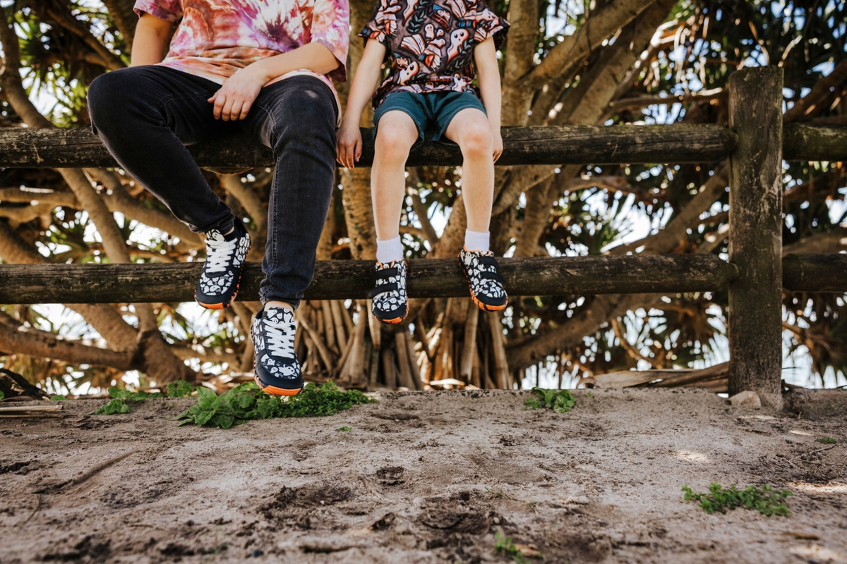 Mum and child sit on a fence at a beach wearing matching PaperKrane ghost print barefoot sneakers. Mum's pair has flat wide white cotton laces, and her child's pair has black velcro straps. Both pairs have orange and black tiger striped soles. 