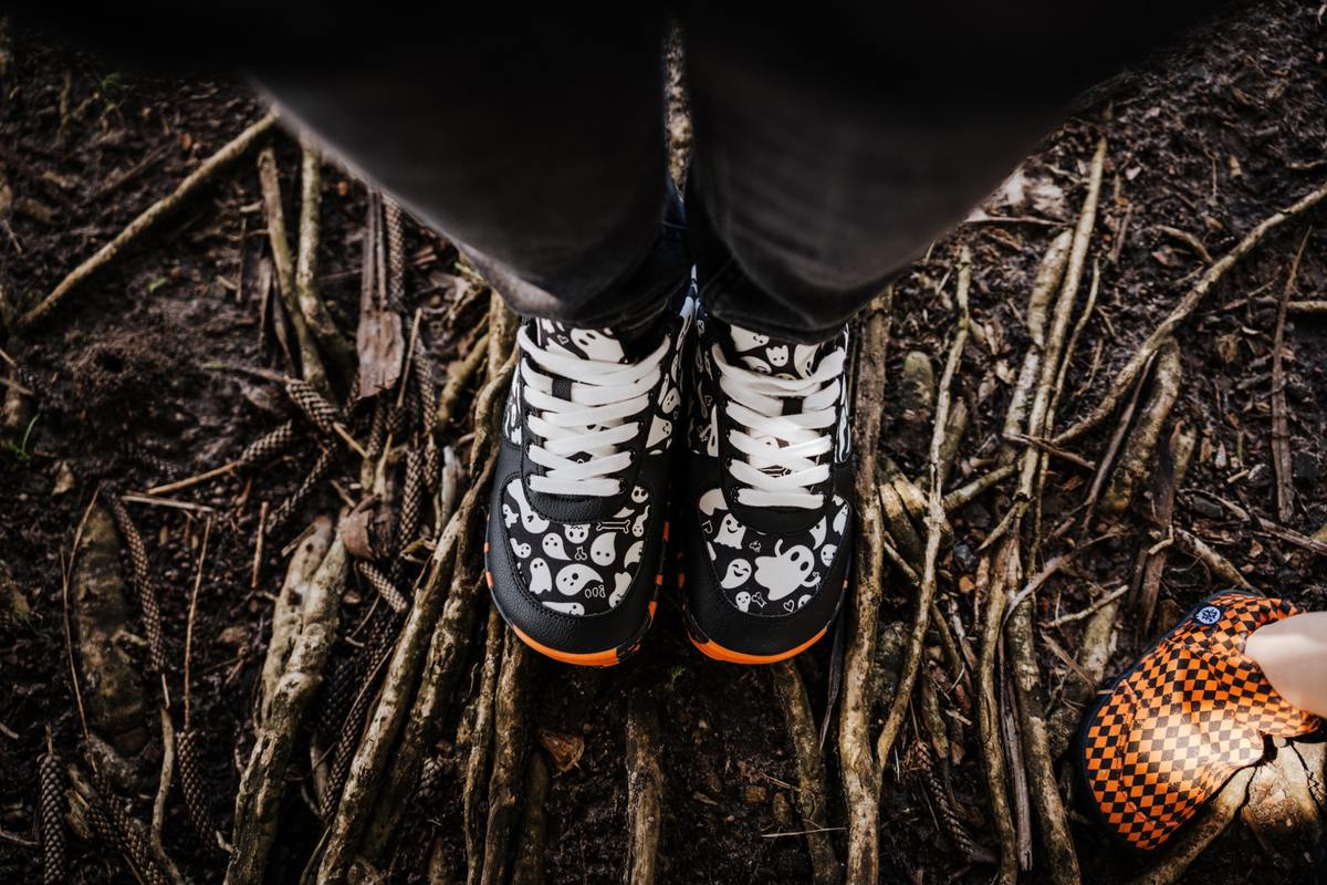 Top down view of Spooky Sneaks, foot-shaped barefoot shoes from PaperKrane. The shoe has a black and white ghost print on an orange and black rubber sole. The person wears black pants and stands on a dirt surface. 