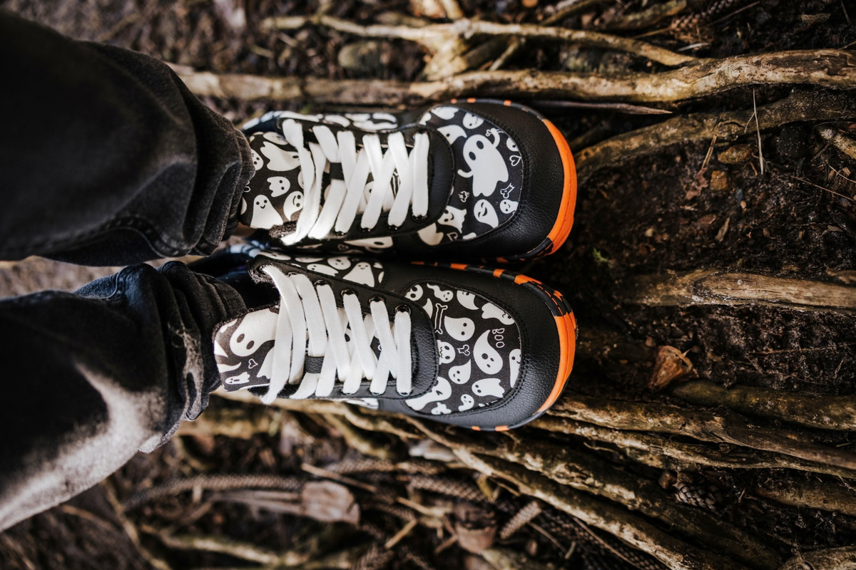 Black and white ghost print PaperKrane barefoot shoes with orange and black rubber soles. They're viewed from above. They're on some wet dirt/tree roots. 