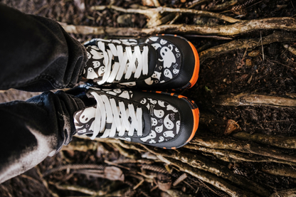Black and white ghost print PaperKrane barefoot shoes with orange and black rubber soles. They're viewed from above. They're on some wet dirt/tree roots. 