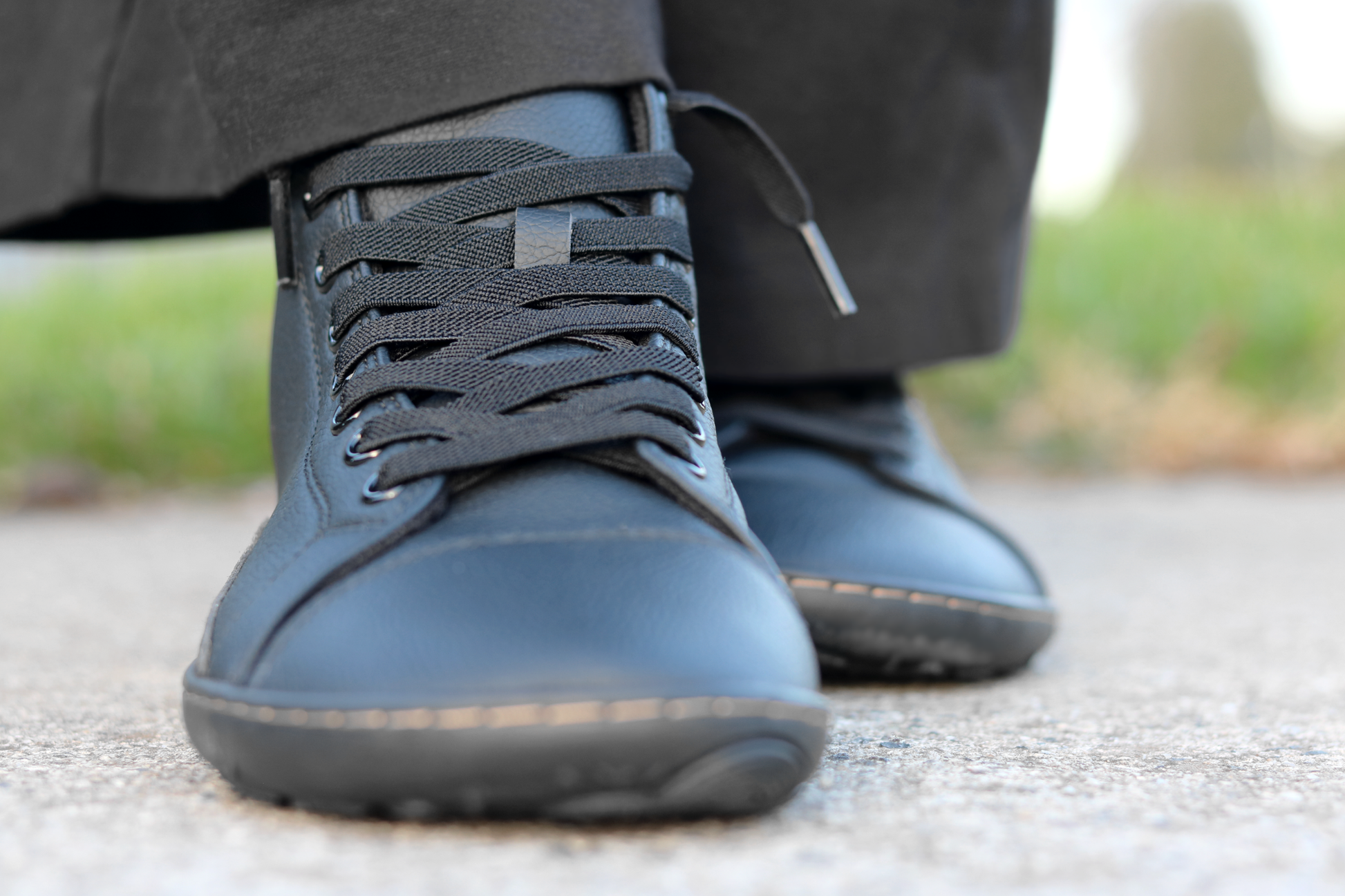 A close-up shot of a person wearing black vegan leather boots with elastic laces, black trousers, standing on a paved outdoor surface.