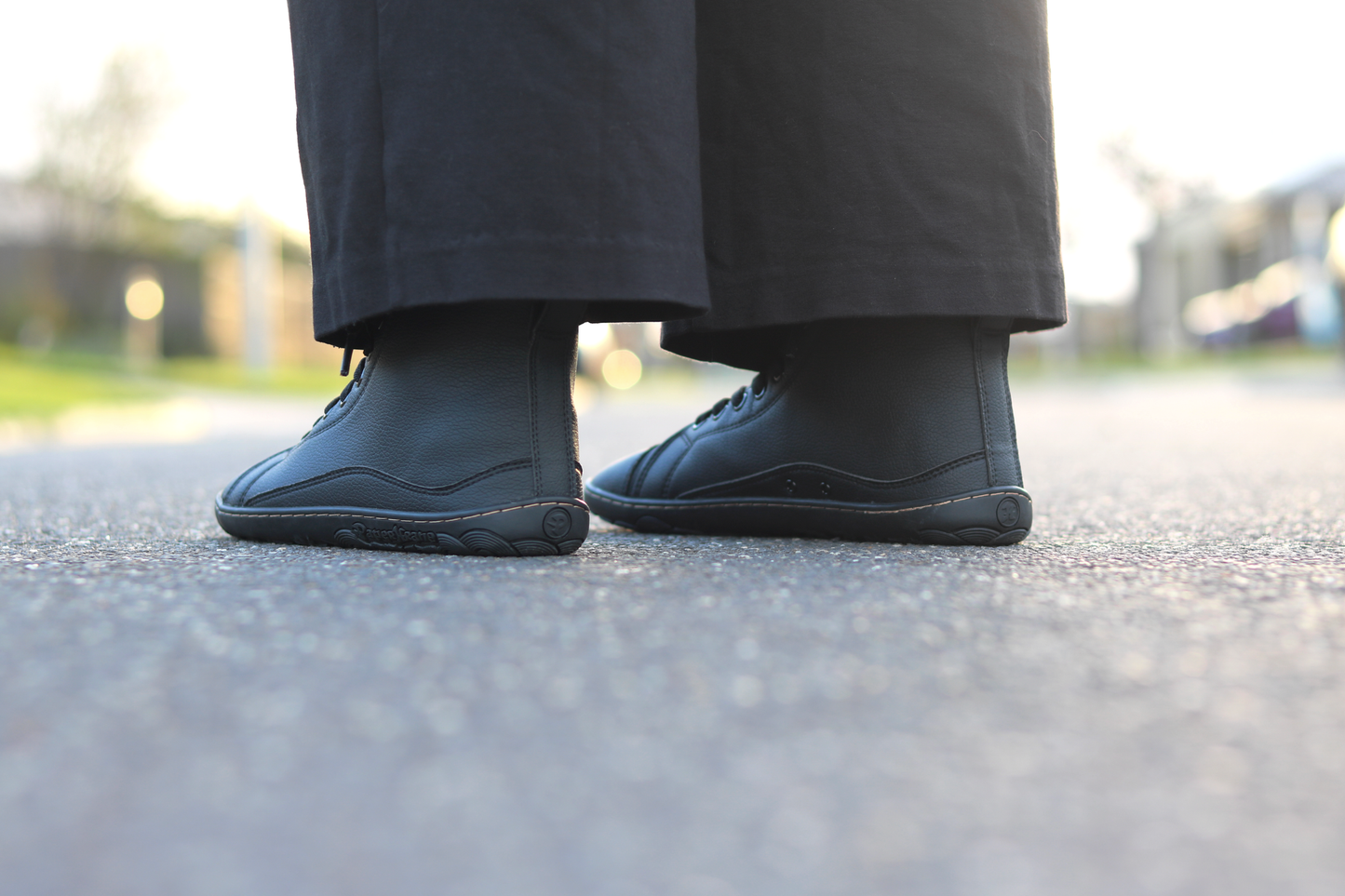 A close-up low-angle shot of a person's feet wearing black vegan leather high-top sneakers and black pants, standing on an asphalt surface with a blurred outdoor background.