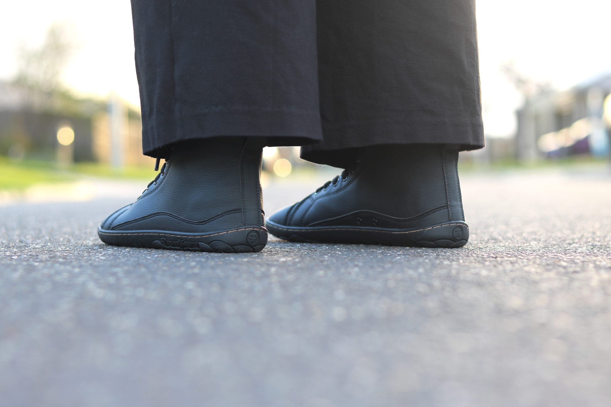 A close-up low-angle shot of a person's feet wearing black vegan leather high-top sneakers and black pants, standing on an asphalt surface with a blurred outdoor background.