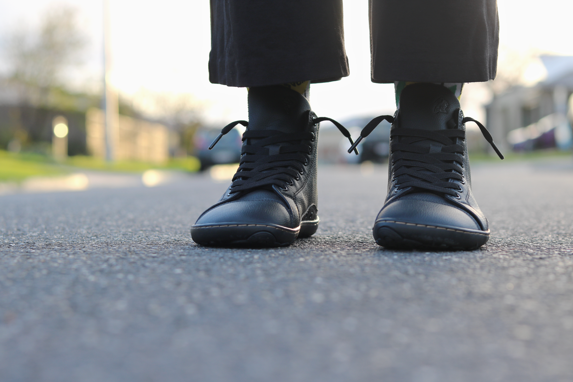 Black vegan leather foot-shaped lace-up ankle boots worn with dark trousers, shown from a low angle on an asphalt street with a blurred outdoor background.