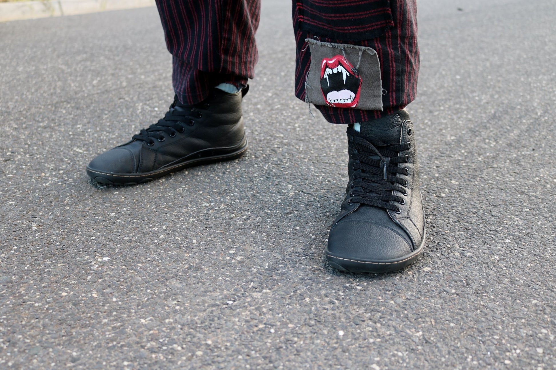 A person standing on gray asphalt wearing black vegan leather barefoot high-top sneakers and dark striped pants with a small red-and-white mouth patch on the lower leg. 