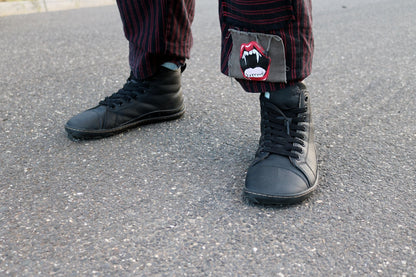 A person standing on gray asphalt wearing black vegan leather barefoot high-top sneakers and dark striped pants with a small red-and-white mouth patch on the lower leg. 
