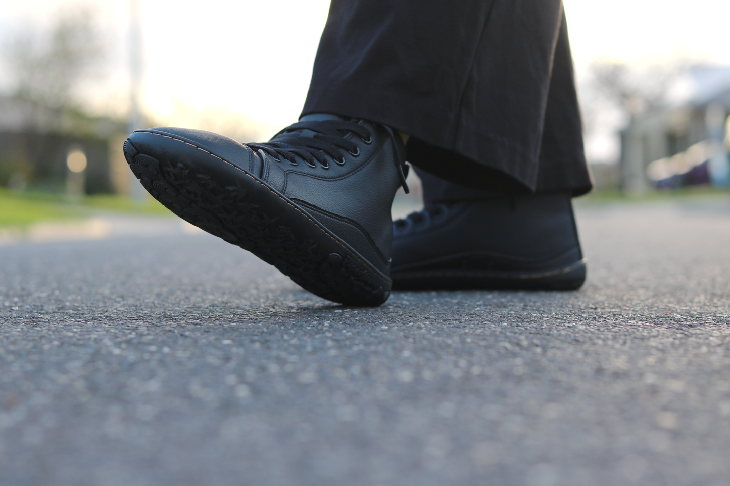 A close-up low-angle shot of a person wearing black vegan leather foot-shaped sneakers and dark trousers, standing on an asphalt road with a blurred outdoor background.