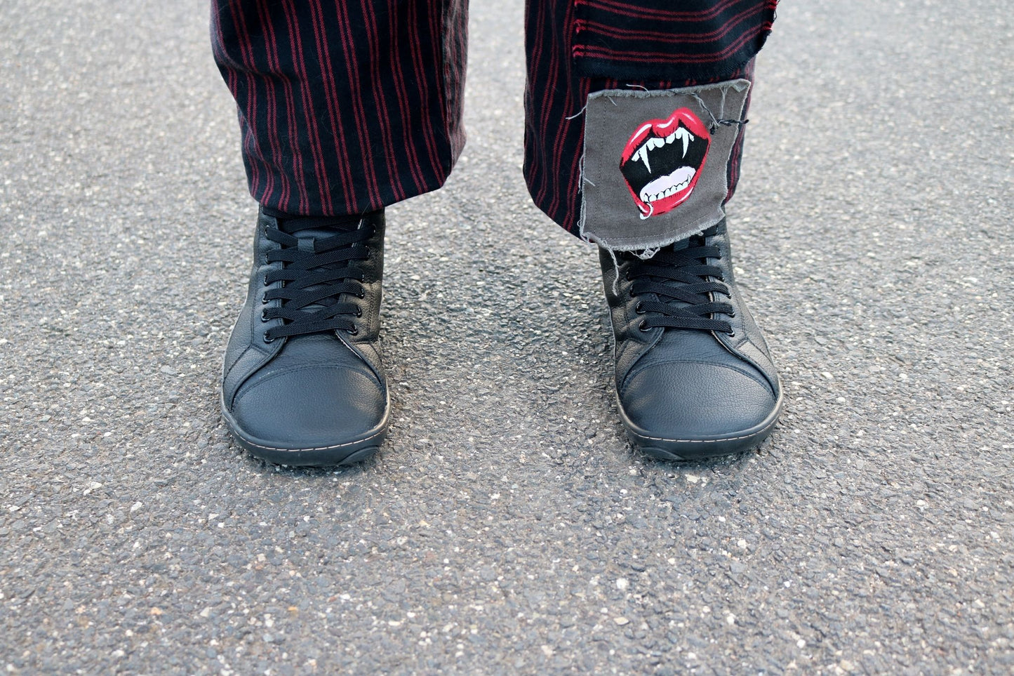 A close-up of a person's feet wearing black vegan leather (elastic) lace-up boots and dark red and black pinstriped trousers, standing on grey asphalt. The right pant leg has a grey patch with a red vampire mouth graphic.