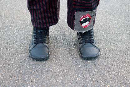 A close-up of a person's feet wearing black vegan leather (elastic) lace-up boots and dark red and black pinstriped trousers, standing on grey asphalt. The right pant leg has a grey patch with a red vampire mouth graphic.