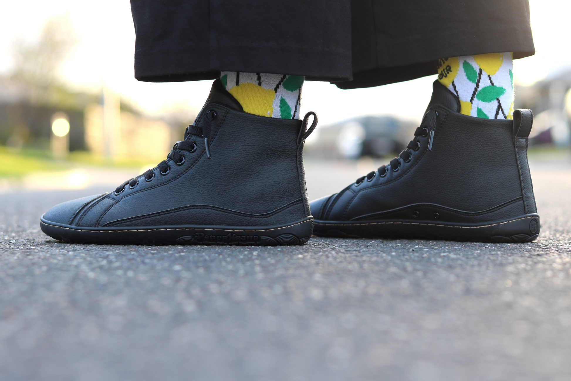A pair of black vegan leather high-top PaperKrane barefoot sneakers worn with colourful yellow, green, and white patterned socks, photographed outdoors on asphalt.