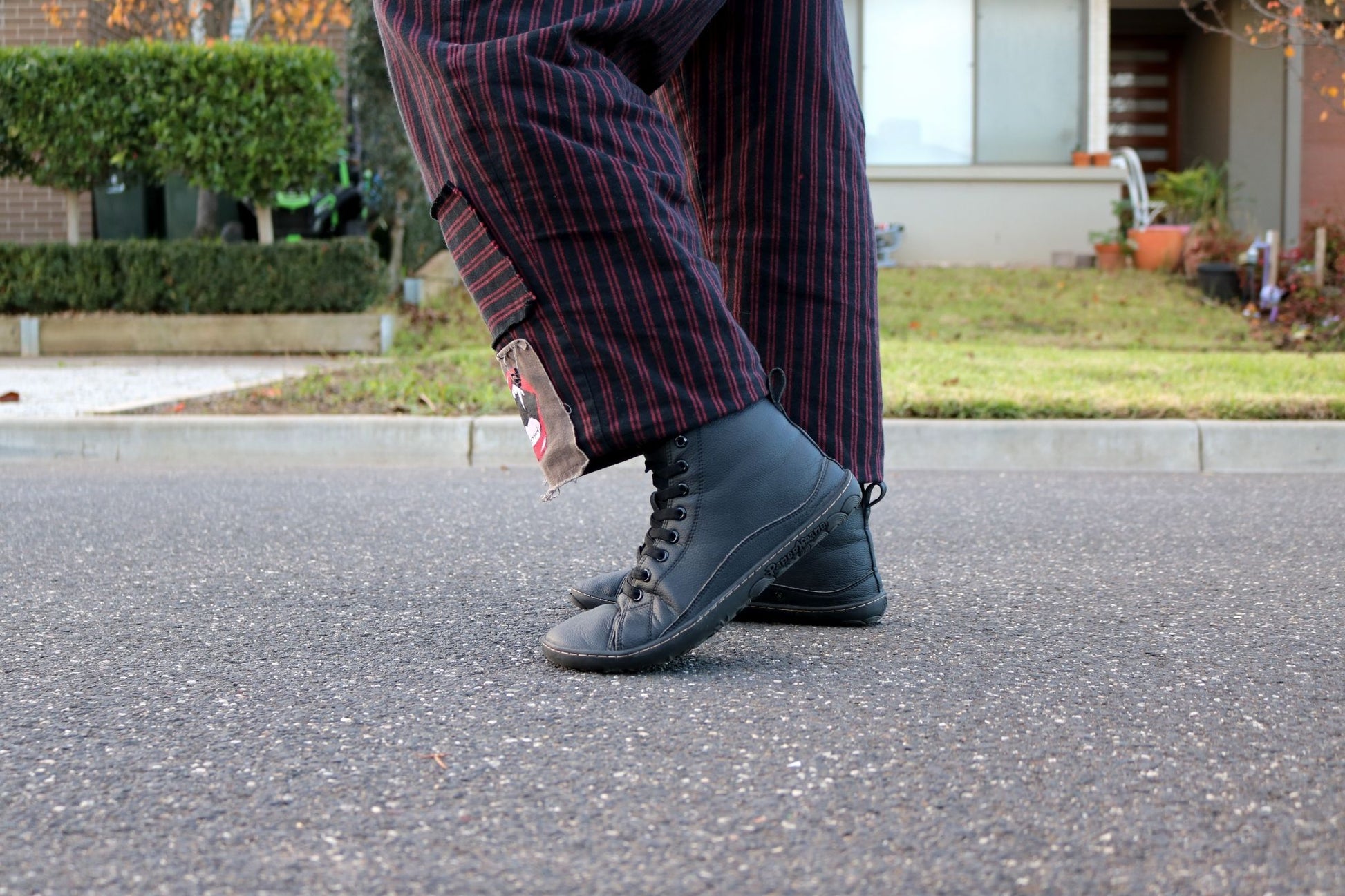 A person wearing black slip on boots with elastic laces and dark red-and-black vertically striped pants, standing on asphalt outdoors with a house and lawn in the background.
