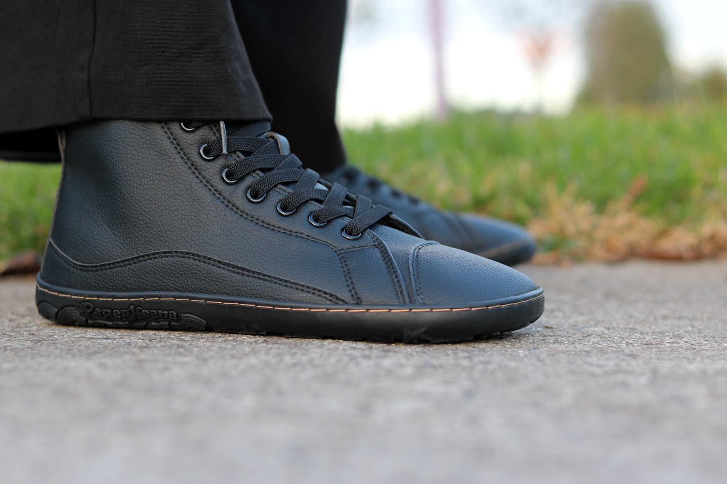 A close-up shot of a person wearing black vegan leather high-top barefoot sneakers outdoors on a paved path with grass in the background. The brand name PaperKrane is embossed on the side of the shoe.