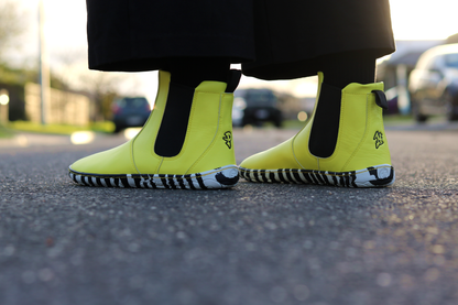 Bright sunny yellow Chelsea-style vegan boots with black and white striped soles, worn with black socks on a blurred street background.