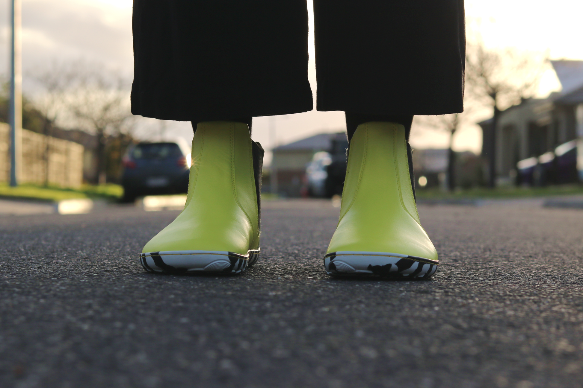Bright sunny yellow Chelsea-style vegan boots from the front with black and white striped soles, worn with black socks on a blurred street background.
