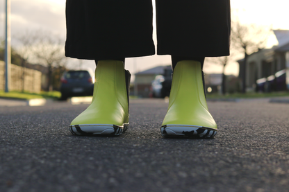 Bright sunny yellow Chelsea-style vegan boots from the front with black and white striped soles, worn with black socks on a blurred street background.