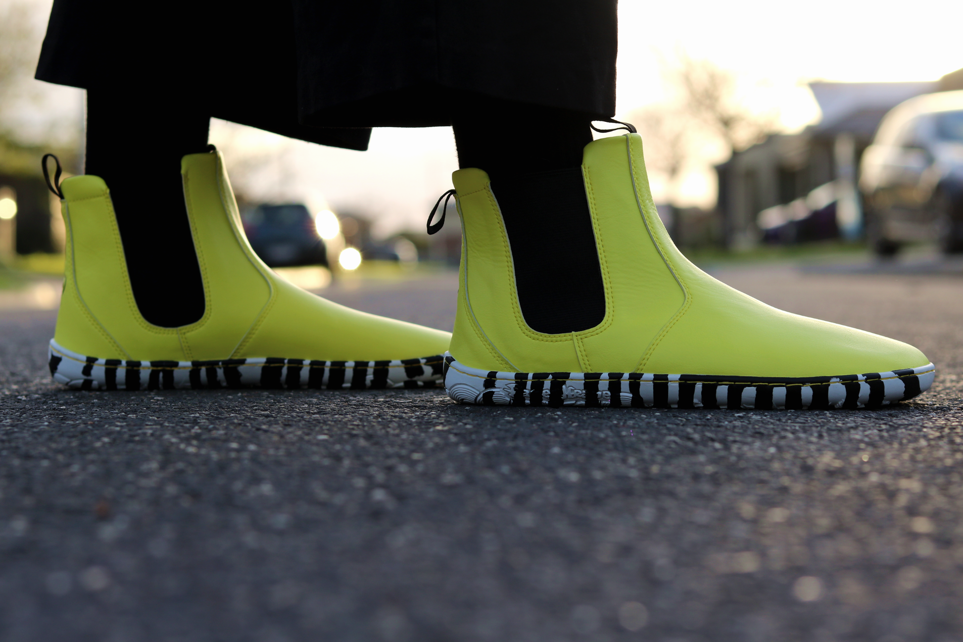 Bright sunny yellow Chelsea-style vegan boots with black and white striped soles, worn with black socks on a blurred street background.