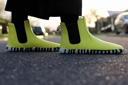 Bright sunny yellow Chelsea-style vegan boots with black and white striped soles, worn with black socks on a blurred street background.