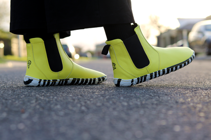 Bright sunny yellow Chelsea-style vegan boots with black and white striped soles, worn with black socks on a blurred street background.