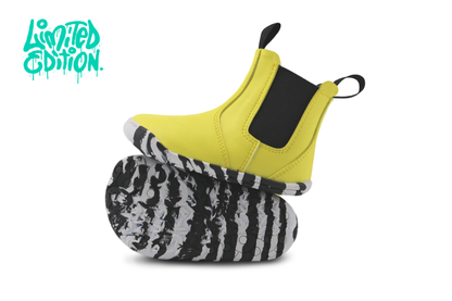 A pair of slip-on toddler chelsea boots shown on a white background, with one boot upright to show the bright yellow upper and black elastic side panel, and the other lying on its side to show a black-and-white zebra-stripe patterned sole. A turquoise 'Limited Edition.