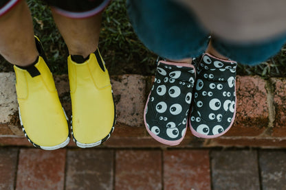 Two foot-shaped slip-on shoes shown from above on a brick ledge outdoors. One shoe is bright yellow with black and white trim, and the other is black with a white eye pattern and salmon pink sole trim.