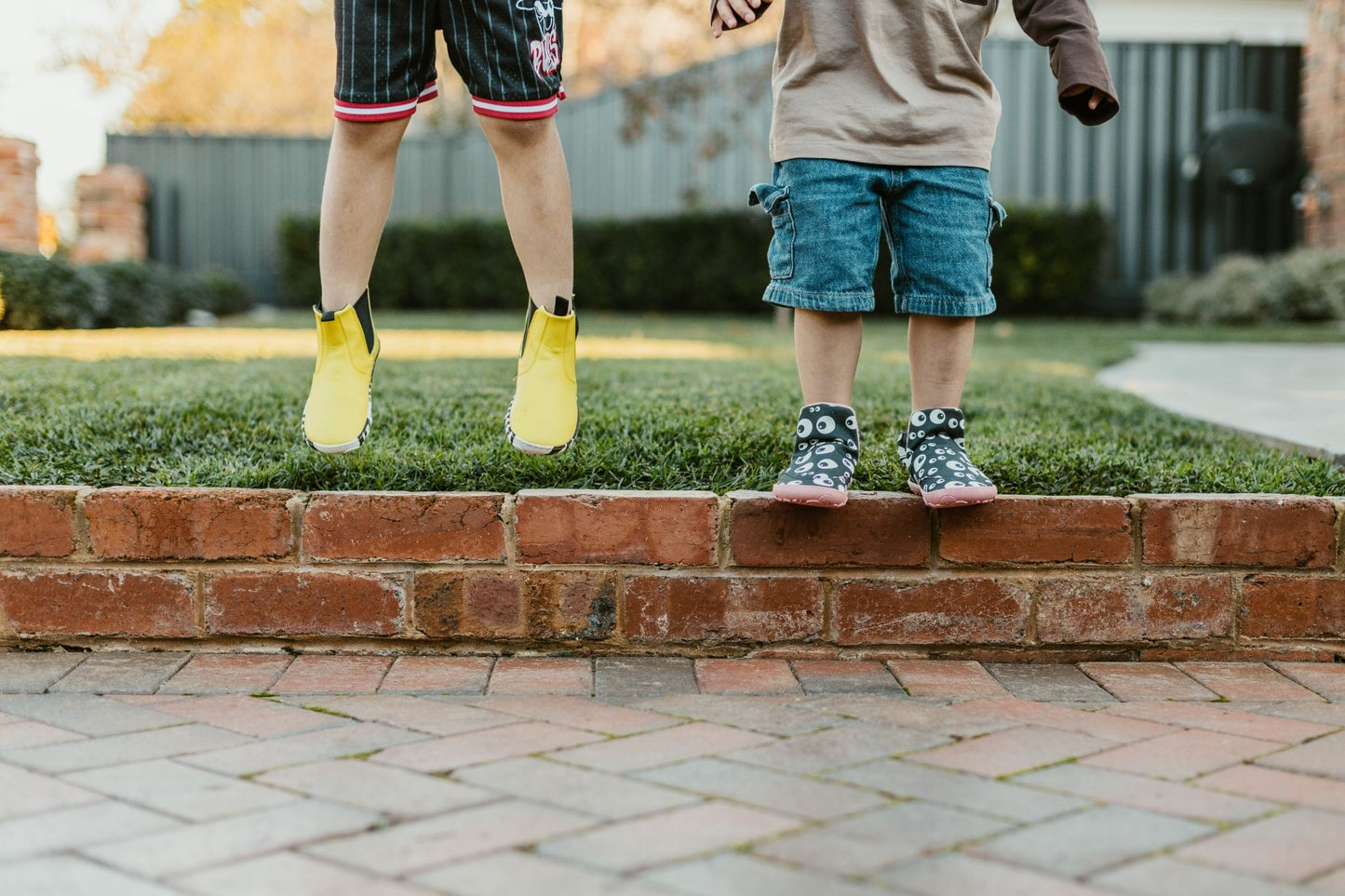 Two children's lower bodies standing on a brick curb in a backyard. One child is jumping in yellow boots, and the other stands in patterned slip on PaperKrane signature style boots.