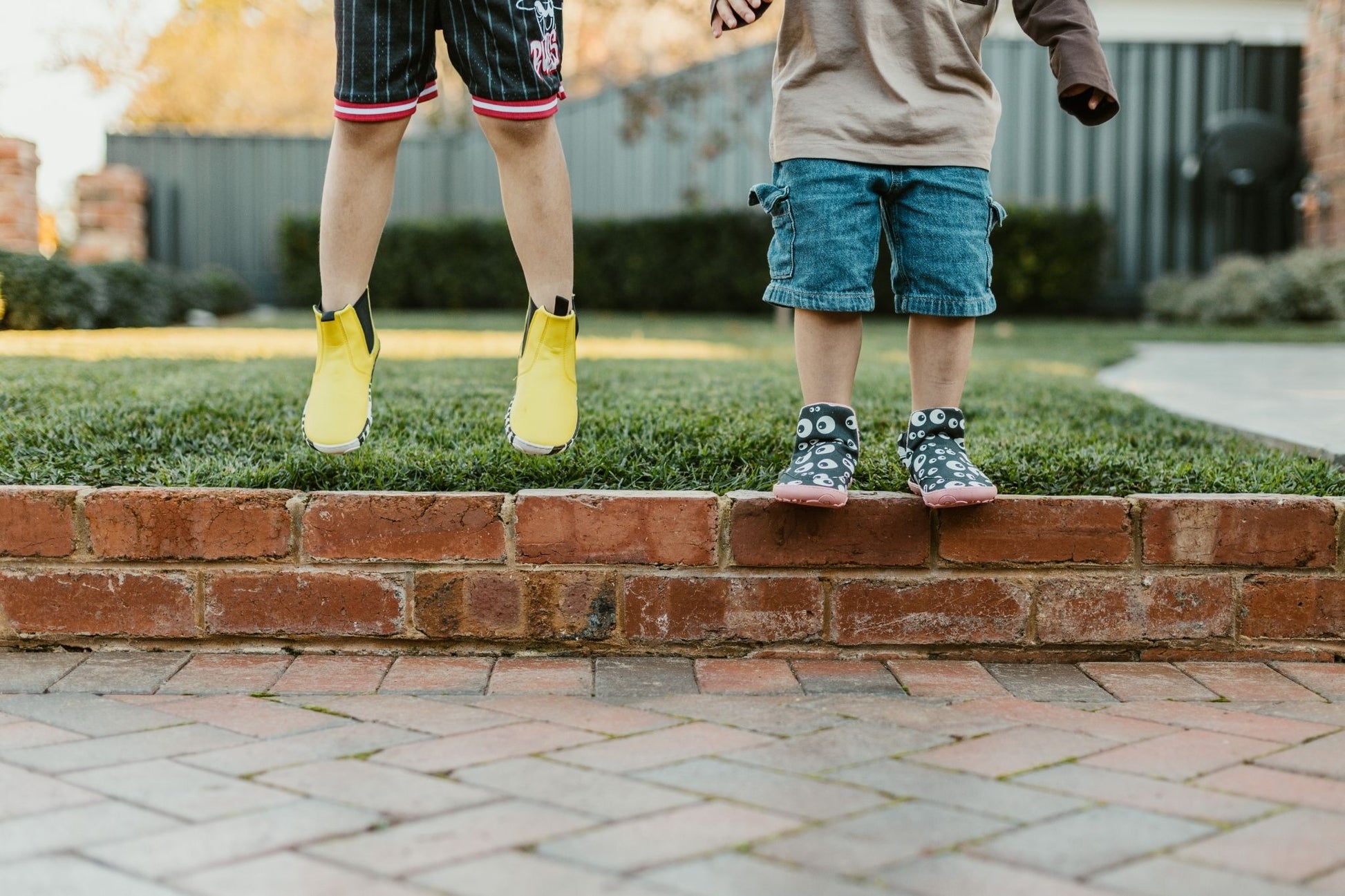 Two children's lower bodies standing on a brick curb in a backyard. One child is jumping in yellow boots, and the other stands in patterned slip on PaperKrane signature style boots.