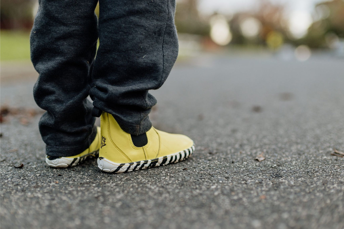 A close-up shot of a child's feet wearing bright yellow slip-on boots with black and white zebra-like soles, paired with dark grey pants, standing on an asphalt surface.