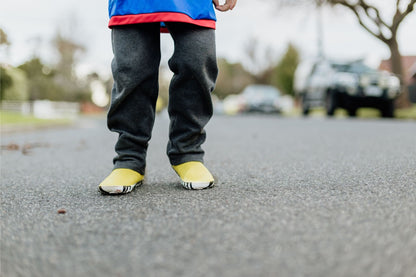 A child's lower body standing on an asphalt road, wearing dark grey pants, a blue and red top, and bright yellow boots.