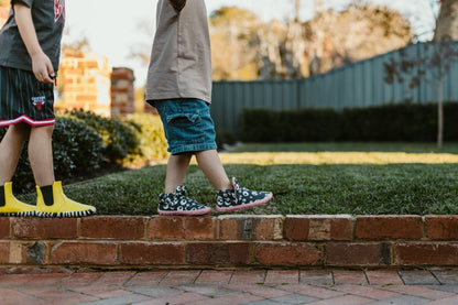 Two children walking outdoors on a brick path beside a grassy yard. One child wears bright yellow barefoot boots and dark shorts, while the other wears patterned slip-on PaperKrane boots with a beige top and denim shorts.